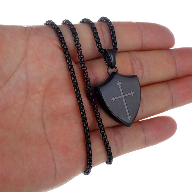 Black shield-shaped pendant with cross design on a chain held in a hand against a white background