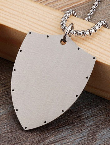 Metallic shield-shaped pendant on a chain, lying on a wooden surface.