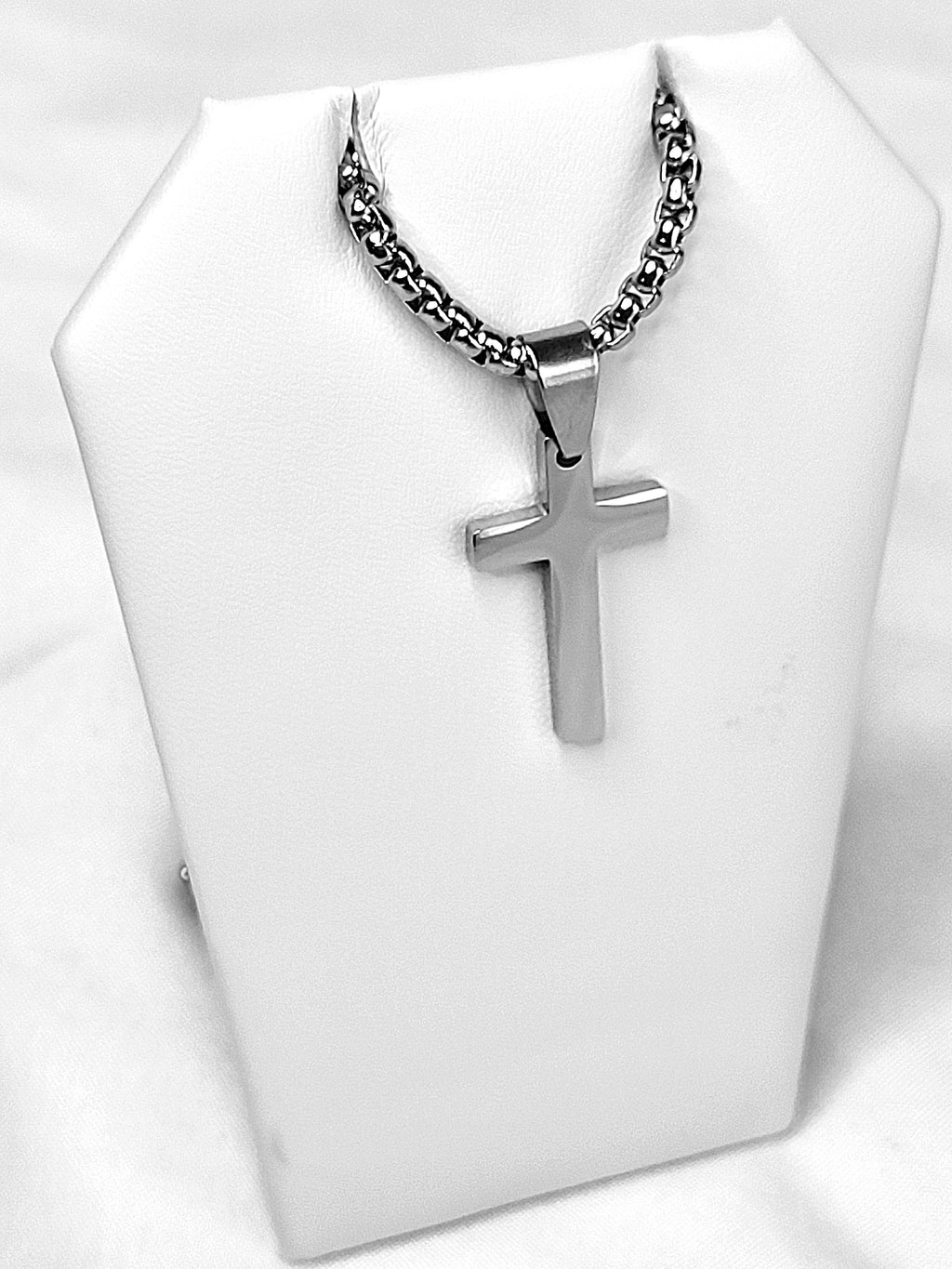 Silver cross pendant on a chain necklace displayed on a white stand.